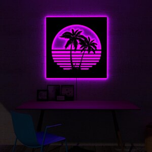 May include: A square, black wall art piece with a neon pink border and a retro sunset design. The design features palm trees, a sun, and horizontal lines. The art is mounted above a desk with a blue chair.