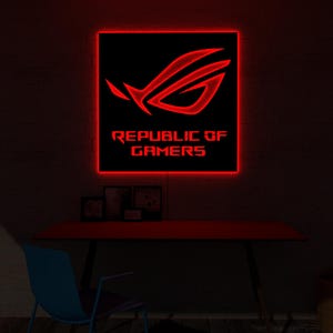 以下が含まれることがあります： 「REPUBLIC OF GAMERS」の文字と、様式化されたロゴが入った赤い照明付きの看板。看板は、赤い机の上にある暗い壁に取り付けられています。机の前には青い椅子があります。