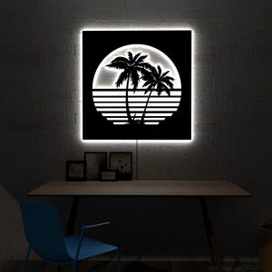 May include: A black and white illuminated wall art piece featuring a tropical sunset scene with palm trees and a setting sun. The artwork is square and has a bright white border. The art is displayed above a wooden desk.