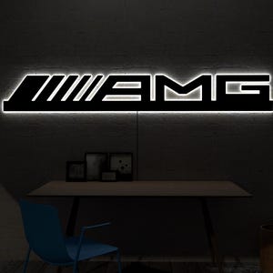 May include: A black AMG logo sign with a white neon backlight. The sign is mounted on a dark gray wall above a wooden desk. A blue chair and a trash can are in front of the desk.