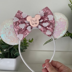 May include: A white headband with sequined mouse ears and a sequined bow. The bow is pink with white heart-shaped sequins. A pink Mickey Mouse-shaped emblem in the centre reads "Bride to be".