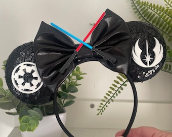 Könnte beinhalten: Schwarzes, paillettenbesetztes Mausohren-Stirnband mit einer großen schwarzen Schleife. Die Ohren zeigen weiße Star Wars-Symbole. Blaue und rote Lichtschwert-Akzente sind an der Schleife befestigt. Ein lustiges Accessoire für Star Wars-Fans.