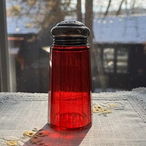 May include: A vintage red glass salt shaker with a silver-colored metal top. The shaker has a ribbed design and is sitting on a white embroidered tablecloth. The background shows a window with a view of trees and a snowy landscape.