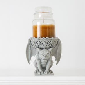 Pode incluir: Um casti&ccedil;al de g&aacute;rgula cinzento com uma vela de frasco de vidro transparente. A vela &eacute; preenchida com cera castanha. A g&aacute;rgula tem asas e est&aacute; sentada. Um elemento decorativo para a casa.