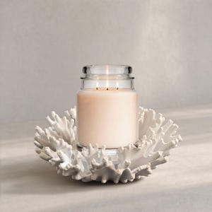 May include: A large, three-wick candle in a clear glass jar, resting on a white coral-shaped holder. The candle has a soft, peach hue and is lit, with three flames visible. The holder is intricately detailed.