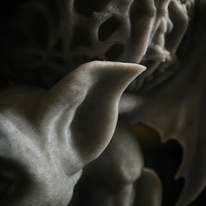 Pode incluir: Close-up de uma escultura texturizada cinza claro sobre um fundo escuro. A escultura tem uma forma de asa e uma orelha pontuda. A superf&iacute;cie &eacute; salpicada com pequenos pontos escuros, adicionando detalhes &agrave; obra.