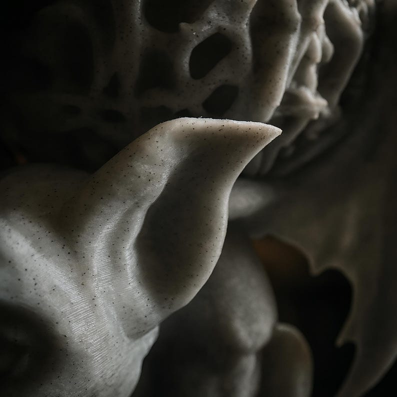 Pode incluir: Close-up de uma escultura texturizada cinza claro sobre um fundo escuro. A escultura tem uma forma de asa e uma orelha pontuda. A superf&iacute;cie &eacute; salpicada com pequenos pontos escuros, adicionando detalhes &agrave; obra.