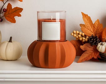 May include: An orange pumpkin-shaped candle holder with a glass-encased candle. The candle has a white label and a lit wick. The background features autumn leaves and small pumpkins, creating a seasonal display.