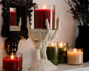 May include: A Halloween-themed candle holder shaped like a skeleton hand, holding a red candle in a glass jar. Other candles in glass jars are in the background, along with a decorative mirror and a black vase with branches.