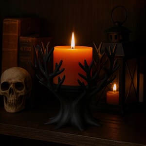May include: A lit orange pillar candle sits in a black, tree-branch-design candle holder. The candle holder is on a dark wooden surface, with a skull and books in the background. A lantern and another candle are also visible.