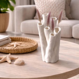Peut inclure: Un vase blanc en forme de corail avec plusieurs ouvertures, contenant plusieurs tiges d'herbe ornementale rose. Le vase est posé sur une table en bois clair, à côté d'un plateau tressé avec une étoile de mer et un coquillage.