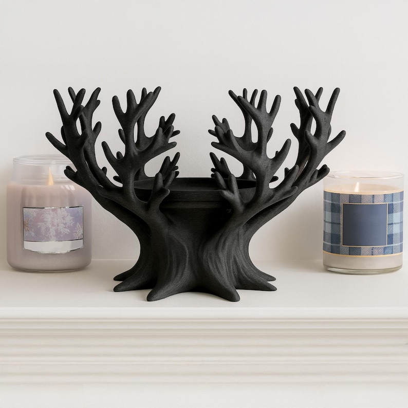May include: A black, tree-shaped candle holder with a central platform, flanked by two lit glass jar candles. The candles have labels and are set on a white shelf. The tree-shaped holder has a textured finish.