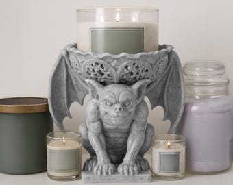 May include: A gray gargoyle candle holder with a large candle and two smaller candles. The candles are in glass jars with labels. A green and gold candle and a purple candle are also present.