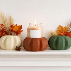 Puede incluir: Tres portavelas con forma de calabaza en crema, óxido y verde. Una vela encendida en un tarro de cristal se encuentra en el soporte de color óxido. El follaje otoñal y el anís estrellado se suman al tema otoñal. Los portavelas están sobre una estantería blanca.