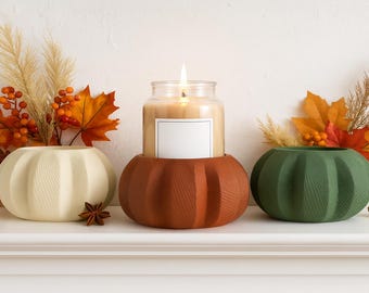 May include: Three pumpkin-shaped candle holders in cream, rust, and green. A lit candle in a glass jar sits in the rust-coloured holder. Autumn foliage and a star anise add to the autumn theme. The holders are on a white shelf.