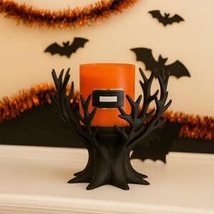 May include: A black, tree-shaped candle holder with an orange candle inside. The candle holder has a decorative design and is set against a Halloween-themed background with bat cutouts and orange garland.