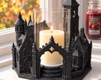 May include: A black candle holder shaped like a gothic cathedral with a central glass cylinder containing a lit cream-colored pillar candle. The structure features intricate architectural details including pointed arches, towers, and stone-like textures.