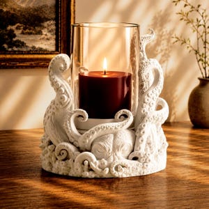 May include: White octopus-themed candle holder with a glass cylinder. A burgundy candle is lit inside. The holder is intricately detailed with tentacles and a central octopus face. The candle holder sits on a wooden surface.