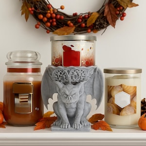 Pode incluir: Tr&ecirc;s velas perfumadas em potes de vidro, uma com r&oacute;tulo vermelho, uma com r&oacute;tulo branco e uma com r&oacute;tulo dourado. Um casti&ccedil;al de g&aacute;rgula cinza suporta uma vela. Uma coroa de outono e folhas est&atilde;o no fundo.