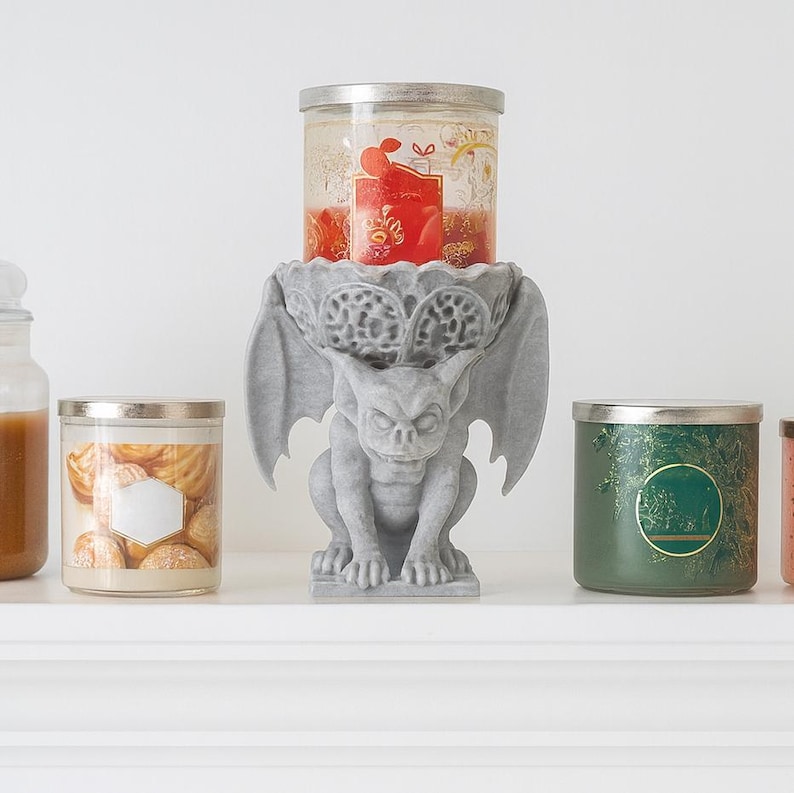 Pode incluir: Um casti&ccedil;al de g&aacute;rgula cinzento com uma vela de vidro. O casti&ccedil;al est&aacute; no centro da imagem, com outras velas de vidro de cada lado. A g&aacute;rgula tem asas e segura a vela.