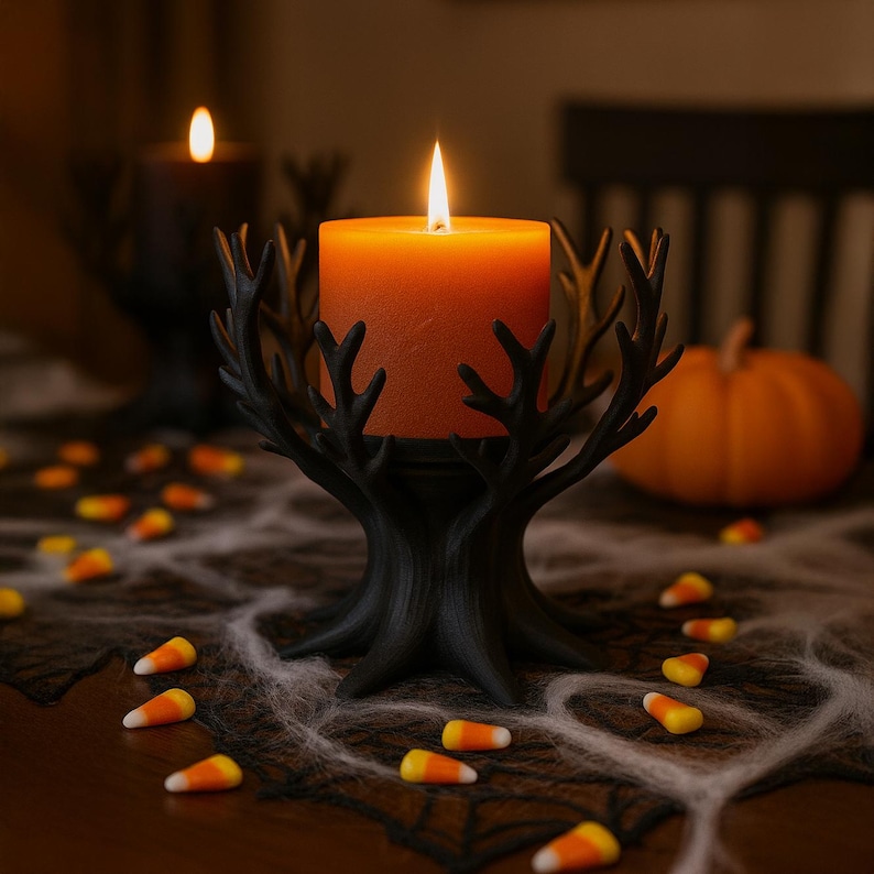 May include: An orange candle, lit, is held in a black tree-shaped candle holder. The candle holder is on a table with a spiderweb runner and candy corn. A small pumpkin is in the background, creating a Halloween theme.