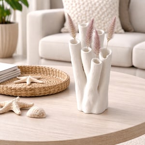 May include: A white ceramic vase with multiple openings, resembling coral, holds dried pink and tan grasses. The vase sits on a light wood table, alongside a woven tray with a starfish and a seashell, creating a coastal theme.
