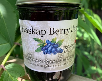 May include: A glass jar of dark purple Haskap Berry Jam with a silver metal lid. The label features a graphic of blue Haskap berries with green leaves, the text "Haskap Berry Jam", "FROM OUR ORCHARD", and "Contains: Mixed haskap berries, sugar, & pectin."