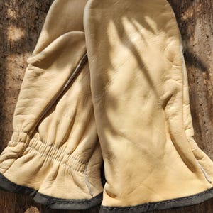 Puede incluir: Un par de guantes de cuero color beige con ribetes negros. Los guantes están hechos de cuero suave y flexible y tienen una sensación cálida y acogedora.