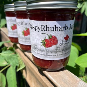 May include: Three jars of "Raspy Rhubarb Jam" with silver lids are displayed. Each jar features a label with a raspberry illustration and the words "Home Grown". The jam inside is a rich, deep red colour, and the jars sit on a wooden surface.