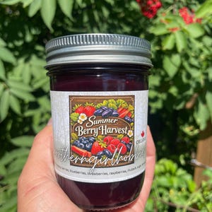 May include: A glass jar filled with dark purple fruit jam, featuring a silver metal lid and a white label. The label displays the text Summer Berry Harvest, an illustration of strawberries, blueberries, and raspberries, and the brand name northerngirlhobbies.