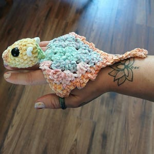 May include: A handmade crochet turtle finger puppet in pastel colors. The turtle's head is yellow and green with a black eye. The shell is a mix of blue, pink, and orange. The puppet is designed to fit on a finger.