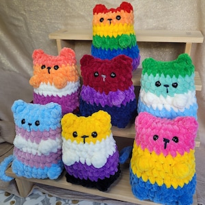 以下が含まれることがあります： 7つの手作りのぬいぐるみ猫のコレクション。各猫は、さまざまなLGBTQ +プライドフラッグを表す異なる配色です。猫は小さな黒いボタンの目とステッチされた鼻を持っています。猫は木製の棚に飾られています。各猫の高さは約15cmです。