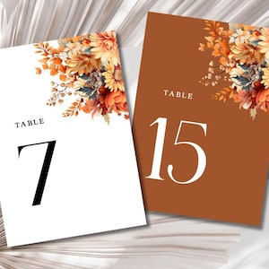 Könnte beinhalten: Zwei weiße Tischkarten mit einem floralen Design in Orange-, Gelb- und Brauntönen. Die Karte links zeigt "TABLE 7" und die Karte rechts zeigt "TABLE 15".