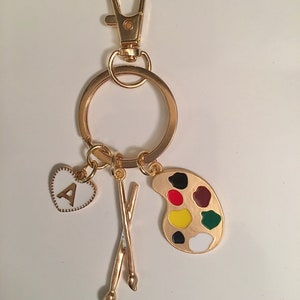May include: Gold-toned keychain with a paint palette charm, paintbrush charm, and a heart charm with the letter A.