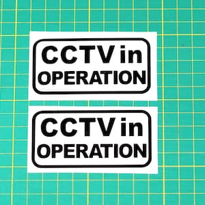 May include: Two white rectangular signs with black borders and black text that reads "CCTV in OPERATION".