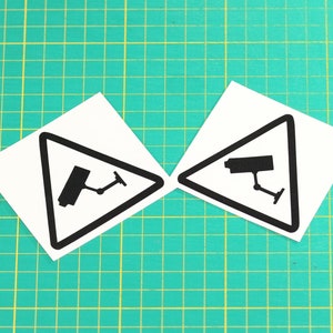 May include: Two white warning signs with black borders and a black silhouette of a security camera inside a triangle. The signs are on a green grid background.