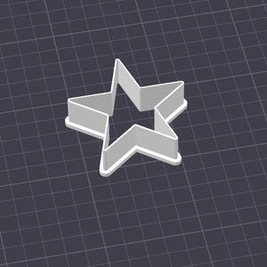 May include: A gray star-shaped cookie cutter with a raised edge. The cutter is made of a light gray material and is set against a dark gray grid background. The star has five points and a hollow center.