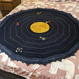 May include: A crocheted blanket in the shape of a solar system with a blue background and a yellow sun in the centre. The planets are represented by different coloured yarn.