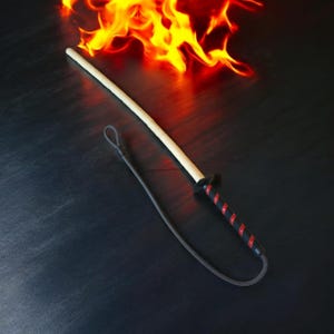 Op de afbeelding: Een trainingszwaard met een lichtgekleurd houten blad en een zwart handvat omwikkeld met een rode spiraalvormige accentlijn. Een zwarte ronde stootplaat scheidt het blad van het handvat, dat een lange zwarte koordlus aan de knop heeft.