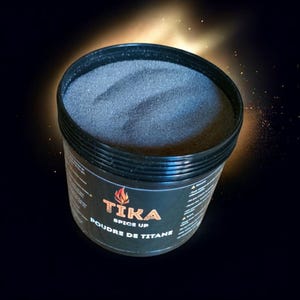 Op de afbeelding: Een zwarte plastic container gevuld met donkergrijs poeder. De container heeft een zwart etiket met de tekst "TIKA SPICE UP POUDRE DE TITANE" in oranje en wit. De achtergrond is donker met een gouden gloed.