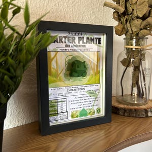 Op de afbeelding: Een ingelijste Pok&eacute;mon-kaart met Bulbasaur, de Grass-type starter Pok&eacute;mon uit de eerste generatie. De kaart is in het Frans en bevat informatie over Bulbasaur's statistieken, evolutie en vaardigheden. De kaart wordt weergegeven tegen een houten achtergrond.