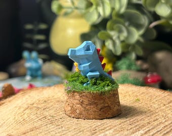Op de afbeelding: Een kleine, lichtblauwe, geometrische dinosaurusfiguur met rode stekels op een miniatuur, met mos bedekte basis. De basis is gemaakt van een cilindrisch, schorsachtig materiaal, geplaatst op een houten schijf.