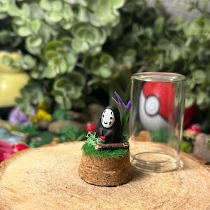Peut inclure: Figurine miniature du personnage Sans-Visage du Voyage de Chihiro, sur une base en liège, à côté d'un petit cylindre en verre. En arrière-plan, un Pokéball flou et de la verdure.