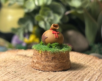 Op de afbeelding: Een miniatuur houten geldbuidel met een rode strik en steraccenten, rustend op een bedje van groen gras en een getextureerde, cilindrische basis. De basis staat op een houten schijf, met een vage groene achtergrond.