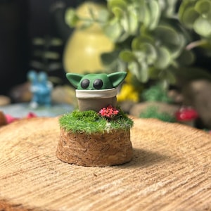 Peut inclure: Figurine artisanale verte et marron représentant un personnage avec de grands yeux noirs et des oreilles pointues. La figurine est posée sur une base en liège avec de la mousse verte et de petites fleurs rouges. L'arrière-plan présente de la verdure.