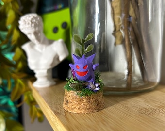 Op de afbeelding: Een paarse, geometrische Gengar-figuur met rode ogen en mond, op een kurken voet met groen mos en kleine paarse bloemen. Het figuur is een decoratief item, mogelijk een verzamelobject of een kleine sculptuur.