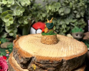 Op de afbeelding: Een kleine, handgemaakte sculptuur van een boomachtig figuur met een hartvormig gezicht, geplaatst op een kleine, cilindrische houten basis. Het figuur is bruin en groen, met een hartvormig gezicht. De achtergrond toont groen en een rood-witte bal.