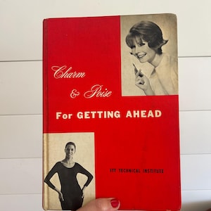 May include: A vintage book titled "Charm & Poise For Getting Ahead" with a red cover. The cover features black and white images of women and the text "ITT TECHNICAL INSTITUTE". The book is held by a person.