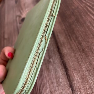 May include: A light green leather wallet with a gold-colored zipper. The wallet is closed and has a rounded rectangular shape. The zipper is partially open, revealing the interior.