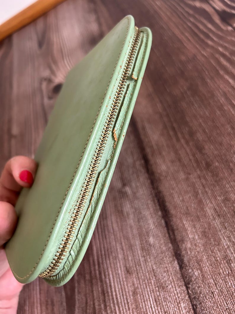 May include: A light green leather wallet with a gold-colored zipper. The wallet is closed and has a rounded rectangular shape. The zipper is partially open, revealing the interior.